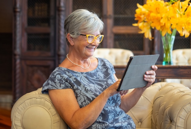 Attraente donna anziana seduta sul divano con tavoletta digitale. Pensionato moderno sorridente che si gode un momento di relax