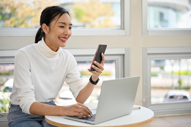 Attraente donna asiatica che usa il suo smartphone mentre lavora a distanza al bar