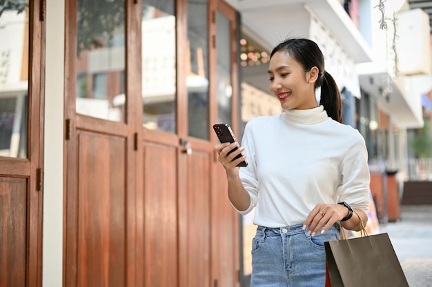 Attraente donna asiatica che utilizza il suo smartphone mentre si diverte a camminare lungo la strada dello shopping