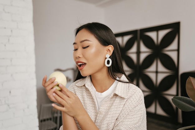 Attraente donna asiatica morde la mela Ritratto di signora di buonumore in cardigan beige e orecchini massicci alla moda tiene frutta fresca