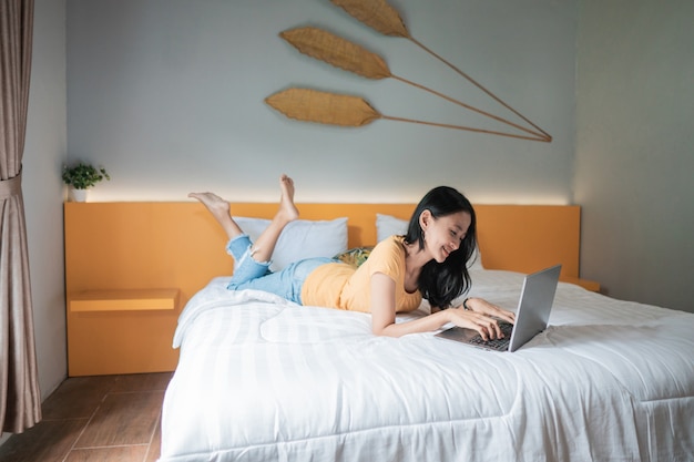 Attraente donna asiatica sdraiata sul letto davanti al suo laptop con le gambe leggermente sollevate