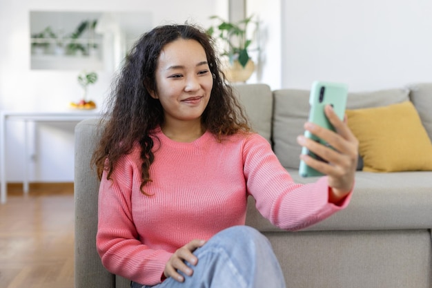 Attraente donna asiatica sorridente che usa lo smartphone mentre si siede sul divano di casa Concetto di comunicazione e videochiamata