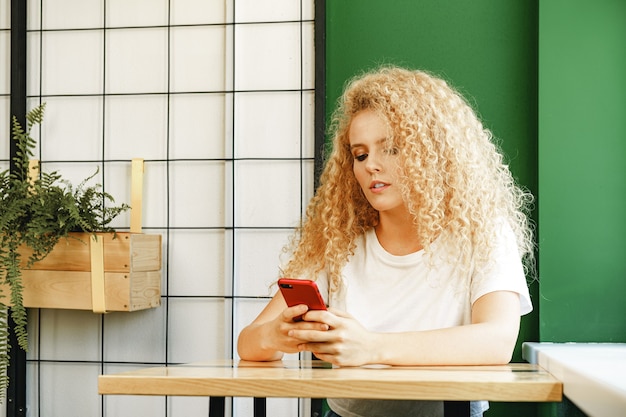 Attraente donna bionda seduta vicino alla finestra nella caffetteria e utilizzando il suo telefono cellulare