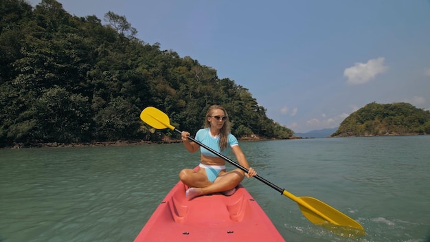 Attraente donna bionda sportiva con occhiali da sole righe rosa canoa di plastica lungo l'acqua di mare contro verdi colline e cielo blu. In viaggio nei paesi tropicali. La ragazza sportiva sta navigando in kayak nell'oceano.