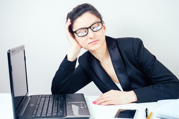 Attraente donna bruna assonnata donna d'affari in elegante tailleur e occhiali con occhi finti dipinti su carta dorme sul posto di lavoro con laptop e un mucchio di cartelle in scadenza di ufficio