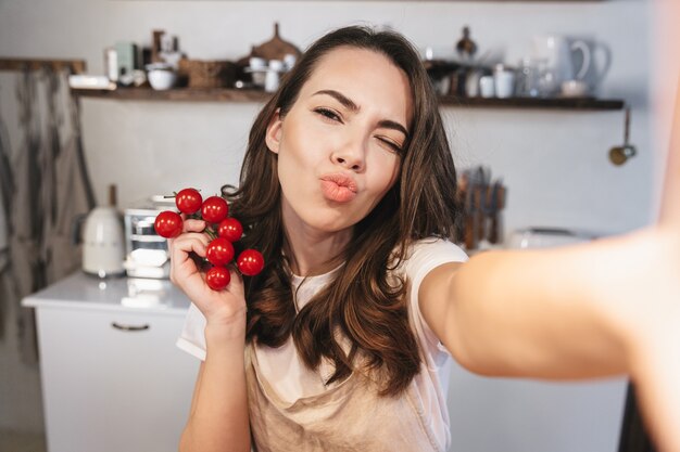 Attraente donna bruna che indossa un grembiule che si fa un selfie mentre è seduta in cucina a casa, con in mano un mazzo di pomodori