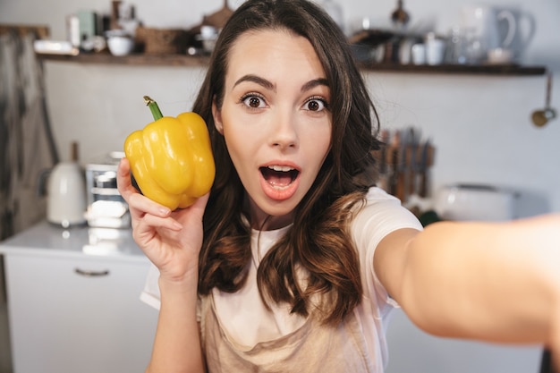 Attraente donna bruna che indossa un grembiule che si fa un selfie mentre è seduta in cucina a casa, con in mano un peperone giallo