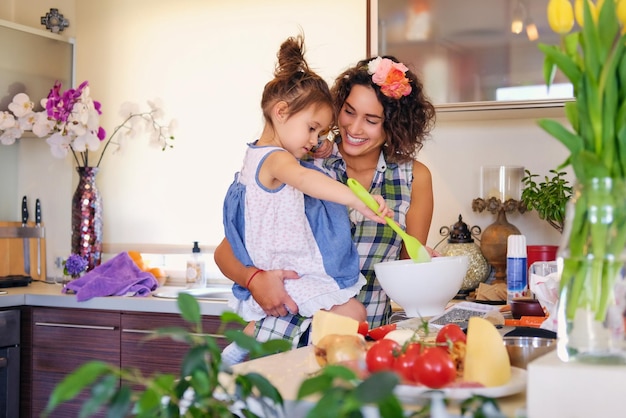 Attraente donna bruna con i capelli ricci e la sua piccola figlia carina che cucinano cibo in una cucina domestica.