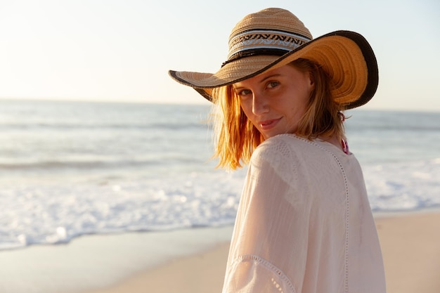 Attraente donna caucasica bionda che si gode il tempo in spiaggia al tramonto, indossa un cappello da sole, guarda l'obbiettivo e sorride, con il cielo al tramonto e il mare sullo sfondo. Vacanze estive al mare tropicale.