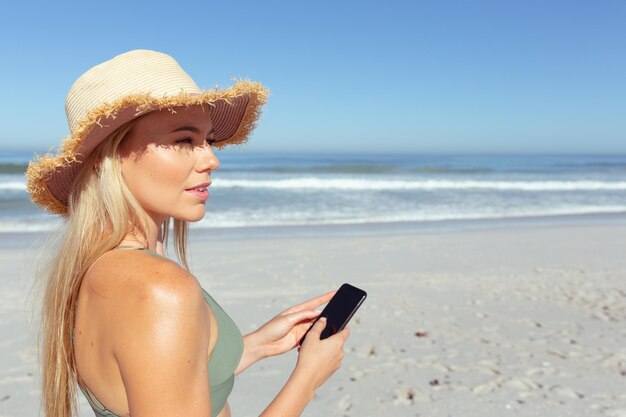 Attraente donna caucasica bionda che si gode il tempo in spiaggia in una giornata di sole, indossa un cappello da sole e utilizza uno smartphone, con cielo blu e mare sullo sfondo. Vacanze estive al mare tropicale.