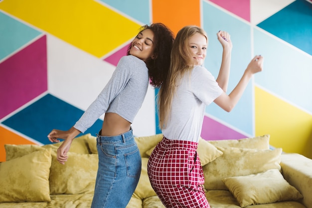 Attraente donna con capelli biondi e bella donna afro-americana che balla con gioia mentre trascorre del tempo insieme al muro colorato