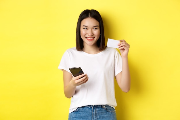 Attraente donna coreana che paga online con lo smartphone, mostrando la carta di credito in plastica e sorridente, in piedi su sfondo giallo.