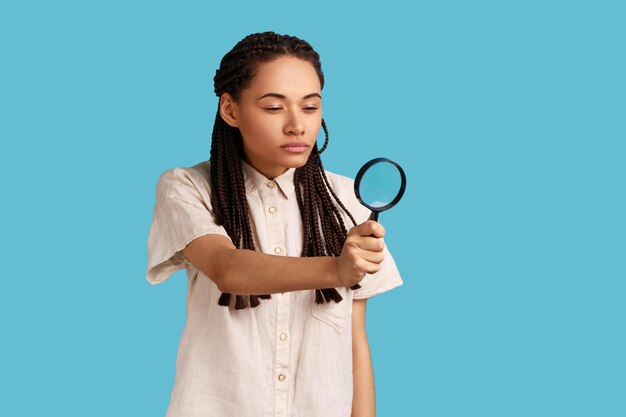 Attraente donna curiosa con i dreadlock neri che distoglie lo sguardo attraverso la lente d'ingrandimento, spia, scopre qualcosa, esplora la scena del crimine, ispeziona. Studio indoor girato isolato su sfondo blu.