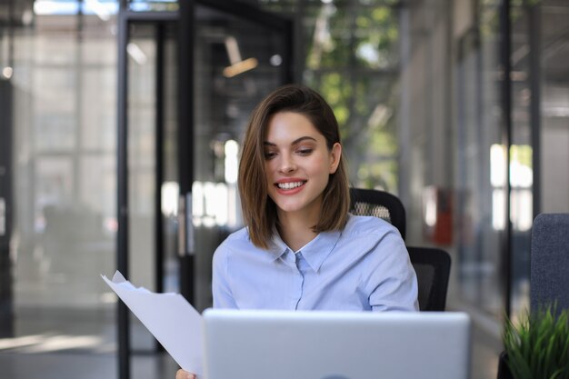 Attraente donna d'affari allegra che controlla i documenti cartacei in ufficio, lavorando sul computer portatile.