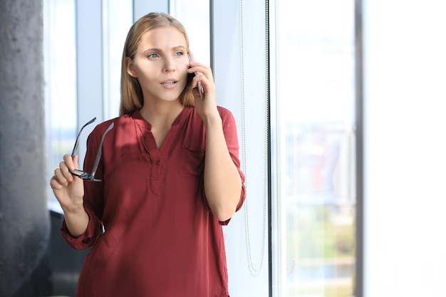 Attraente donna d'affari che parla con i colleghi al telefono cellulare mentre si trova vicino alla finestra in ufficio.