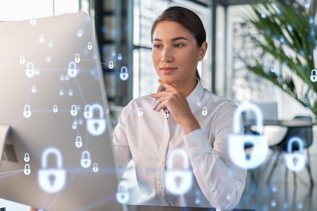 Attraente donna d'affari in camicia bianca sul posto di lavoro che lavora con il laptop per difendere la sicurezza informatica dei clienti Concetto di protezione delle informazioni dei clienti Ologramma del lucchetto sullo sfondo dell'ufficio