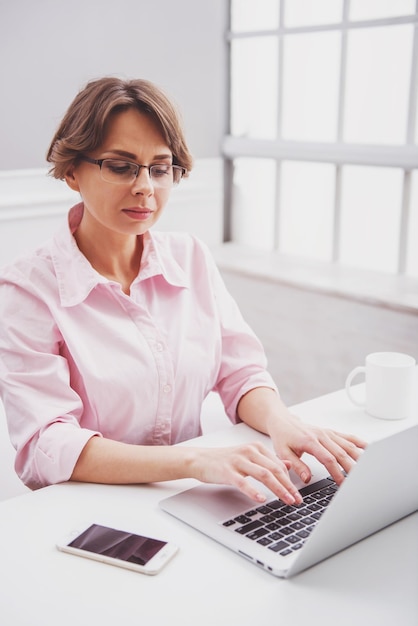 Attraente donna d'affari in camicia classica e occhiali sta usando un laptop mentre lavora in ufficio