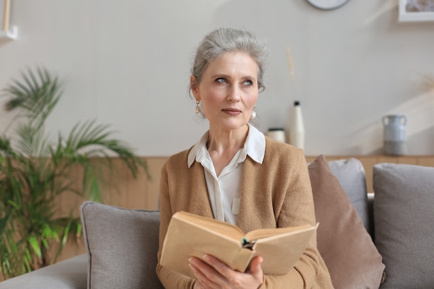 Attraente donna di mezza età che si gode la lettura di un libro seduta sul divano nel suo soggiorno sorridendo mentre legge.