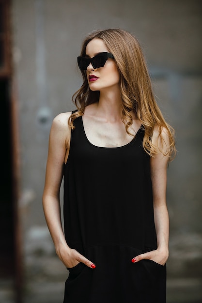 Attraente donna di moda in abito nero con occhiali da sole in posa vicino al muro bianco