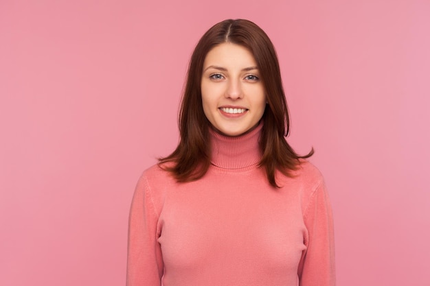 Attraente donna di successo con capelli castani in maglione rosa che guarda l'obbiettivo con un sorriso a trentadue denti, felice soddisfatto della vita, libero professionista. Studio indoor girato isolato su sfondo rosa