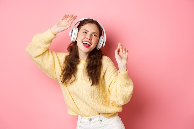 Attraente donna elegante che canta la canzone preferita, ascolta la musica in cuffie wireless, sorride e balla, in piedi sopra il muro rosa.