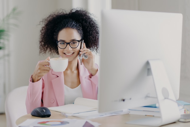 Attraente donna elegante dai capelli ricci beve caffè aromatico, pone sul desktop con computer moderno necessario per il lavoro, chiama amico durante la pausa, discute le ultime notizie, ha un sorriso a trentadue denti