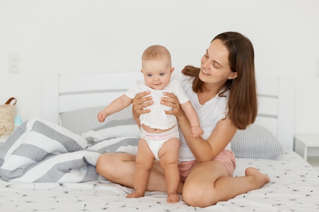 Attraente donna felice con i capelli scuri che indossa una camicia bianca e breve seduta sul letto con sua figlia bambino in piedi con la mamma aiuta la madre a guardare il bambino con un sorriso affascinante