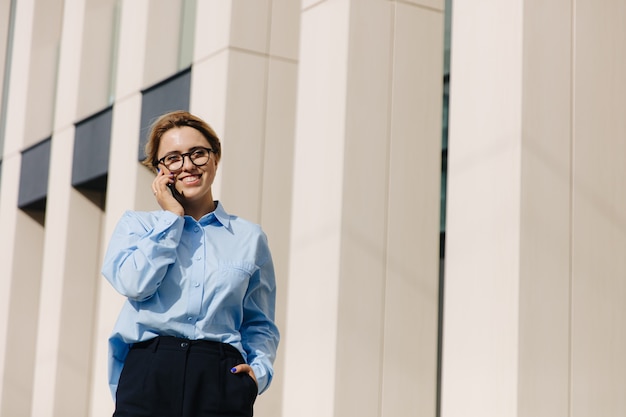 Attraente donna in camicia blu e pantaloni neri utilizzando il cellulare per chattare all'aria aperta. Donna sorridente con capelli biondi che gode della conversazione all'aperto.