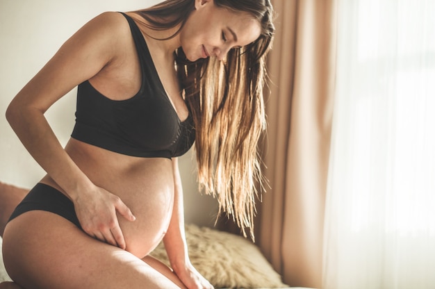 Attraente donna incinta è seduta a letto e si tiene la pancia