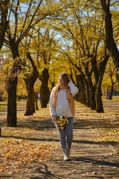 Attraente donna incinta millenaria con fiori nel parco autunnale. Bella futura madre che si diverte all'aperto nella soleggiata giornata autunnale e si gode la bellissima natura all'aperto