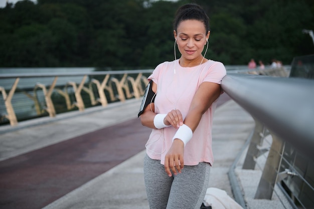 Attraente donna ispanica in cuffia e una maglietta rosa si trova su un ponte della città e indossa braccialetti da tennis in spugna prima del jogging mattutino e dell'allenamento cardio. Bella atleta femminile che si allena all'aperto