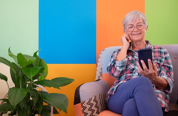 Attraente donna moderna hipster sorridente sedersi comodamente sul divano in soggiorno a casa ascoltare musica con gli auricolari con smart phone in mano Senior donna godere della tecnologia