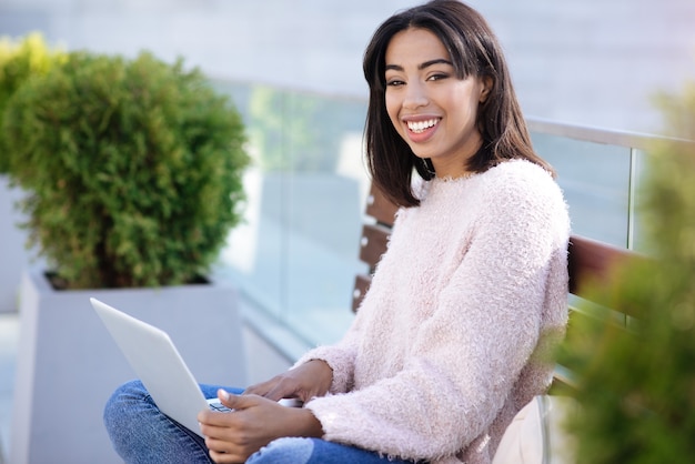 Attraente donna motivata dinamica che si siede nel parco e lavora al suo nuovo progetto mentre prende appunti usando il suo laptop