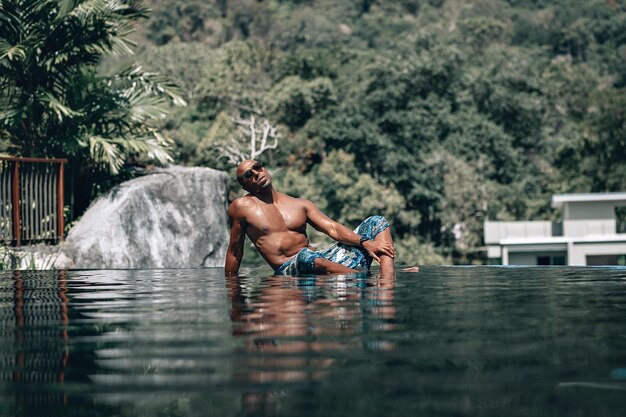 Attraente e rilassato maschio arabo senza camicia in sarong turchese sdraiato sul lato di una piscina a sfioro in una giornata di sole; riflesso nell'acqua, palme e hotel sullo sfondo; concetto di vacanza.