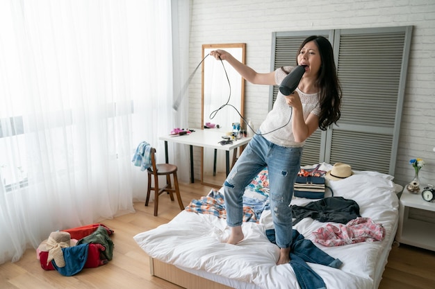 attraente felice giovane donna asiatica in abbigliamento casual che canta all'asciugacapelli sul letto in casa accogliente camera da letto. turismo viaggio d'affari persone e concetto di bagaglio. la borsa da viaggio per l'imballaggio della signora si rilassa divertendosi da sola