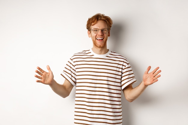 Attraente giovane con i capelli rossi e gli occhiali che mostrano grandi dimensioni, lunghezza di un grande oggetto, sorridendo soddisfatto, in piedi su sfondo bianco.