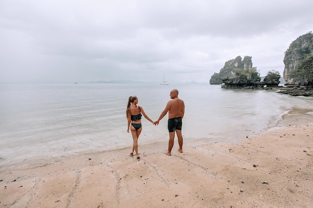 Attraente giovane coppia alla moda in abiti bianchi cammina sulla spiaggia e si tiene per mano. La bella bionda con i capelli lunghi e un uomo calvo sono in vacanza. Phuket. Tailandia. Vista posteriore.