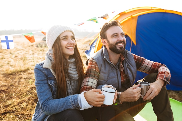 Attraente giovane coppia che riposa mentre è seduto alla tenda all'aperto, tenendo tazza e thermos