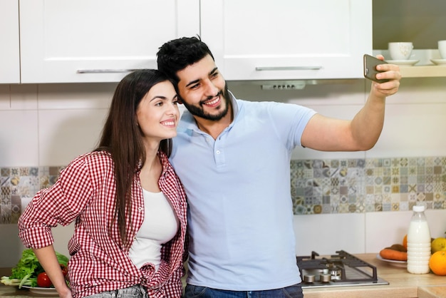 Attraente giovane coppia che si fa selfie con lo smartphone indossando abiti casual alla moda divertendosi insieme nell'accogliente cucina