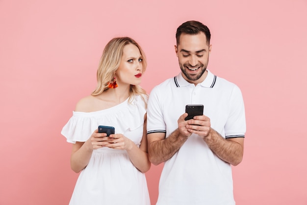 Attraente giovane coppia curiosa che sta insieme isolata sopra il rosa, usando il telefono cellulare