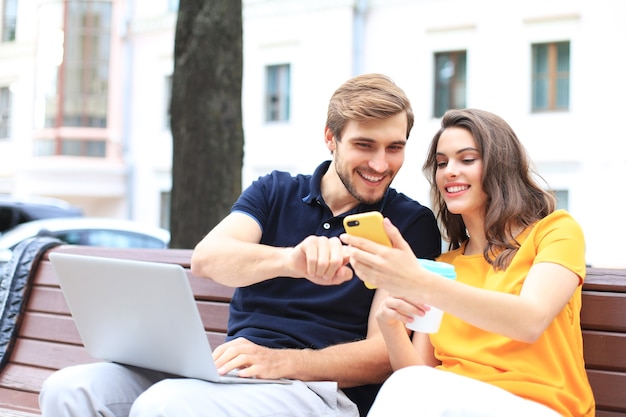 Attraente giovane coppia seduta su una panchina e guardando qualcosa nel telefono cellulare.