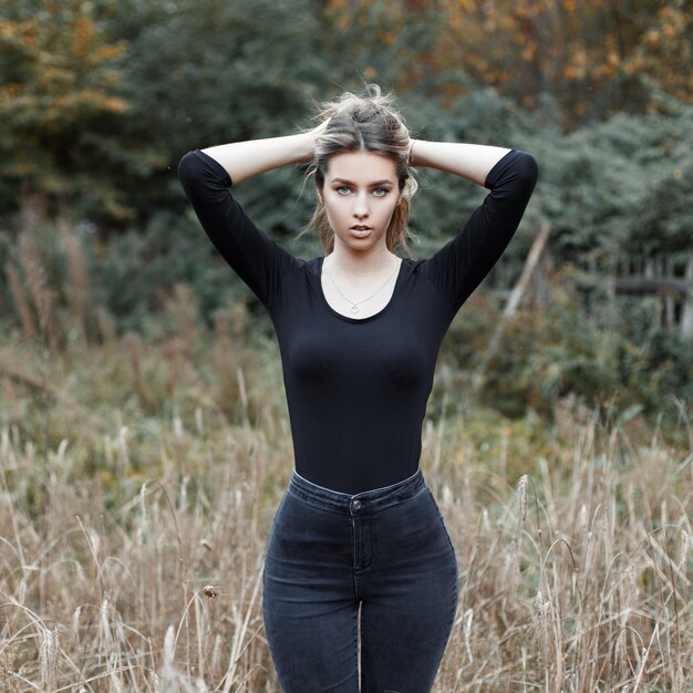 Attraente giovane donna abbastanza bella in un'elegante maglietta in jeans vintage in piedi in un campo tra l'erba secca