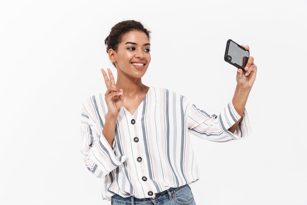 Attraente giovane donna africana che indossa abiti casual in piedi isolato su un muro bianco, facendo un selfie, mostrando pace