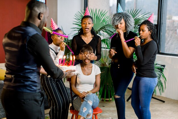 Attraente giovane donna africana con i capelli scuri, seduta sulla sedia con i suoi amici intorno a lei, festeggia e spegne le candeline di compleanno. Un ragazzo africano porta una torta di compleanno con candele