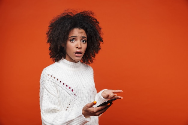 Attraente giovane donna africana confusa che indossa un maglione tenendo il telefono cellulare isolato su un muro rosso