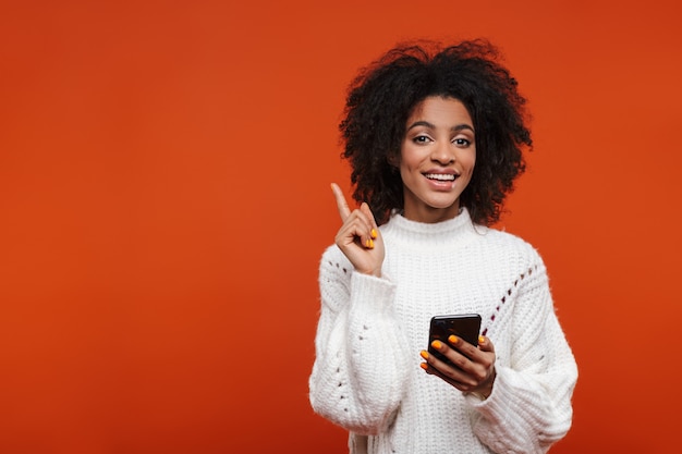 Attraente giovane donna africana sorridente che indossa un maglione che tiene il telefono cellulare isolato sul muro rosso, puntando il dito lontano