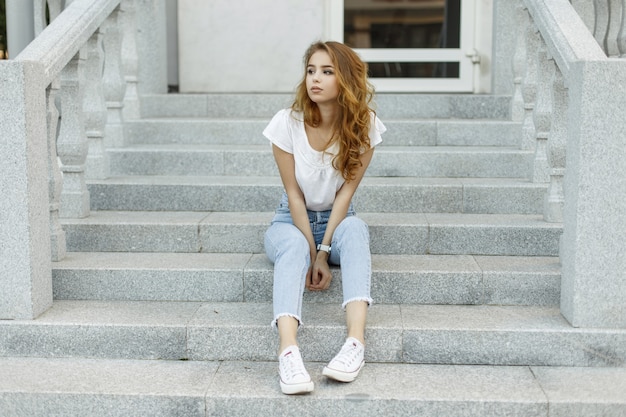 Attraente giovane donna alla moda in una maglietta alla moda in blue jeans in scarpe da ginnastica alla moda bianche che riposa seduto sui gradini di un edificio vintage.