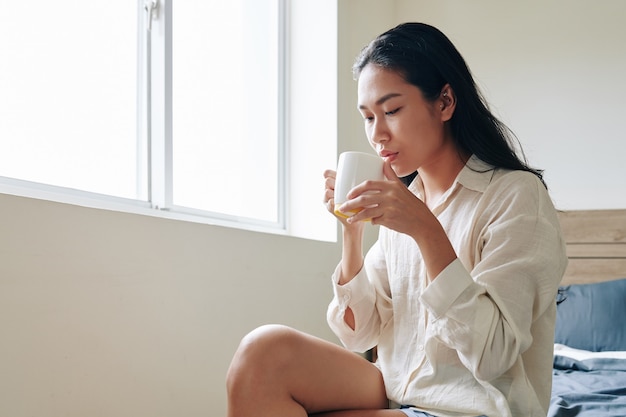 Attraente giovane donna asiatica seduta sul letto e gustando una grande tazza di gustoso caffè al mattino