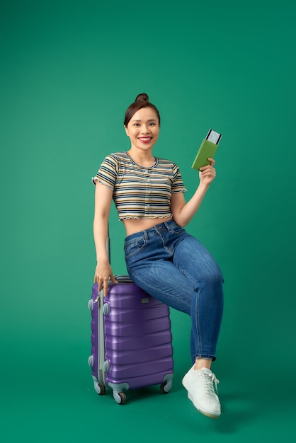 Attraente giovane donna asiatica seduta sulla valigia e tenendo il passaporto, biglietto di volo sul verde.