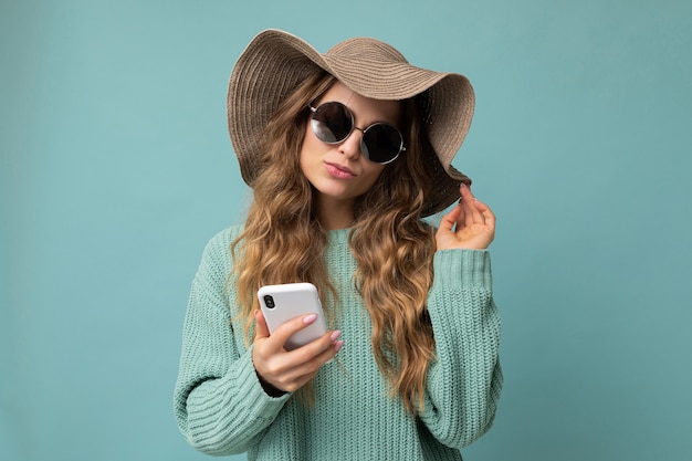 Attraente giovane donna bionda che indossa un maglione blu cappello e occhiali da sole in piedi isolato su blue